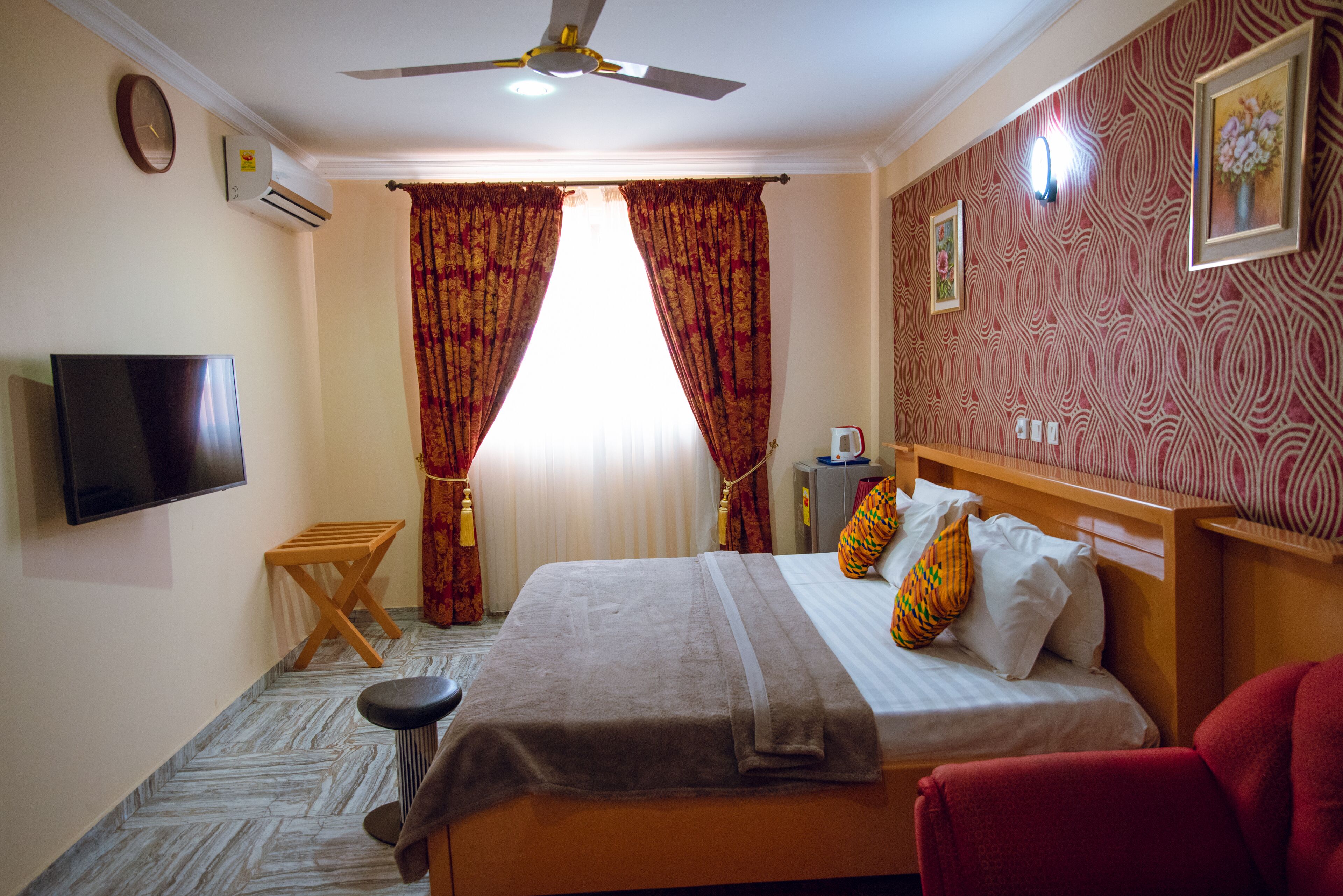Habitación de hotel con cama, televisor, ventilador de techo, una mesita y una silla roja.