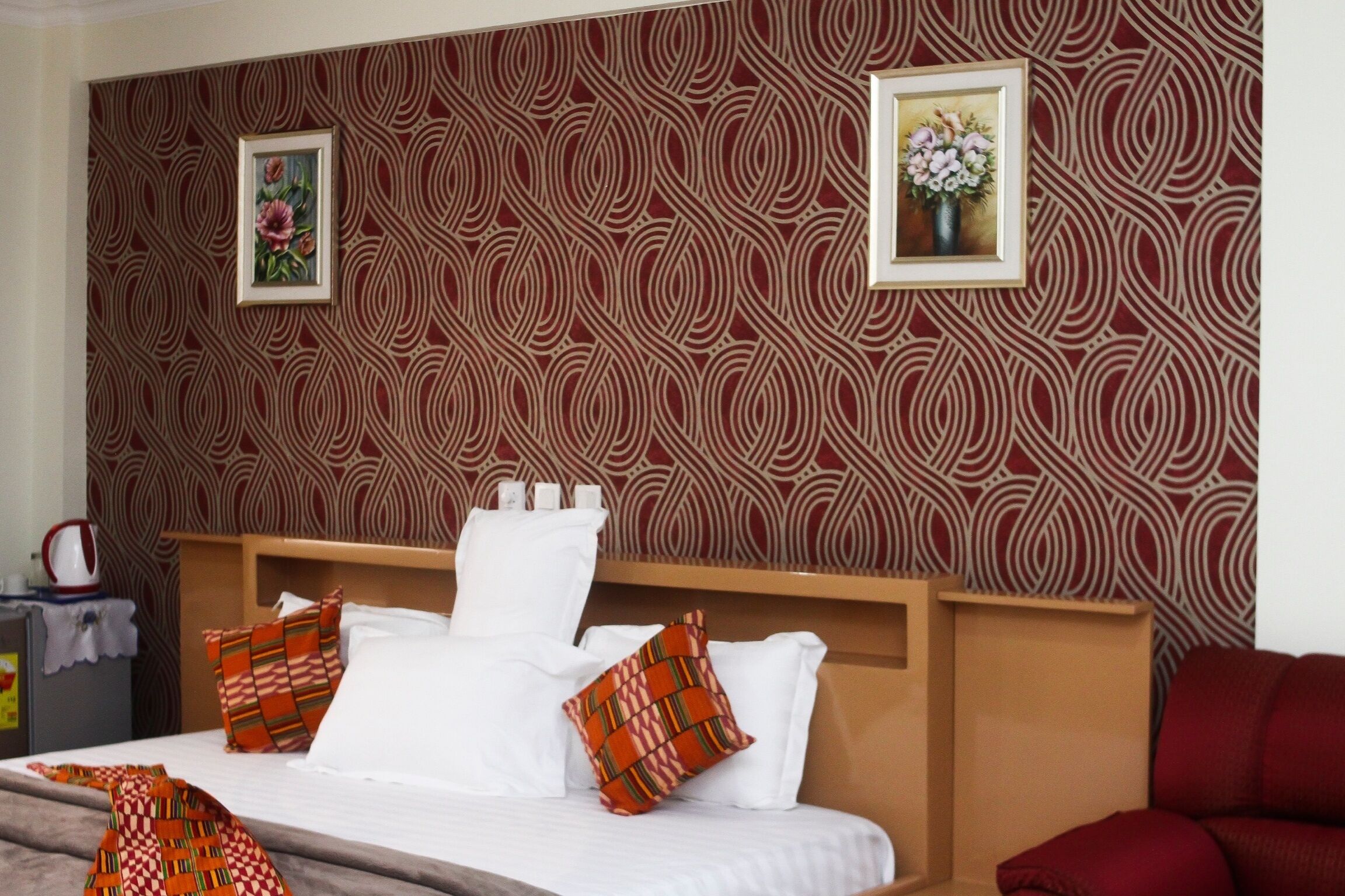 A hotel room with a patterned wallpaper, a bed with white linens and multiple pillows, a wooden headboard, and a red sofa.