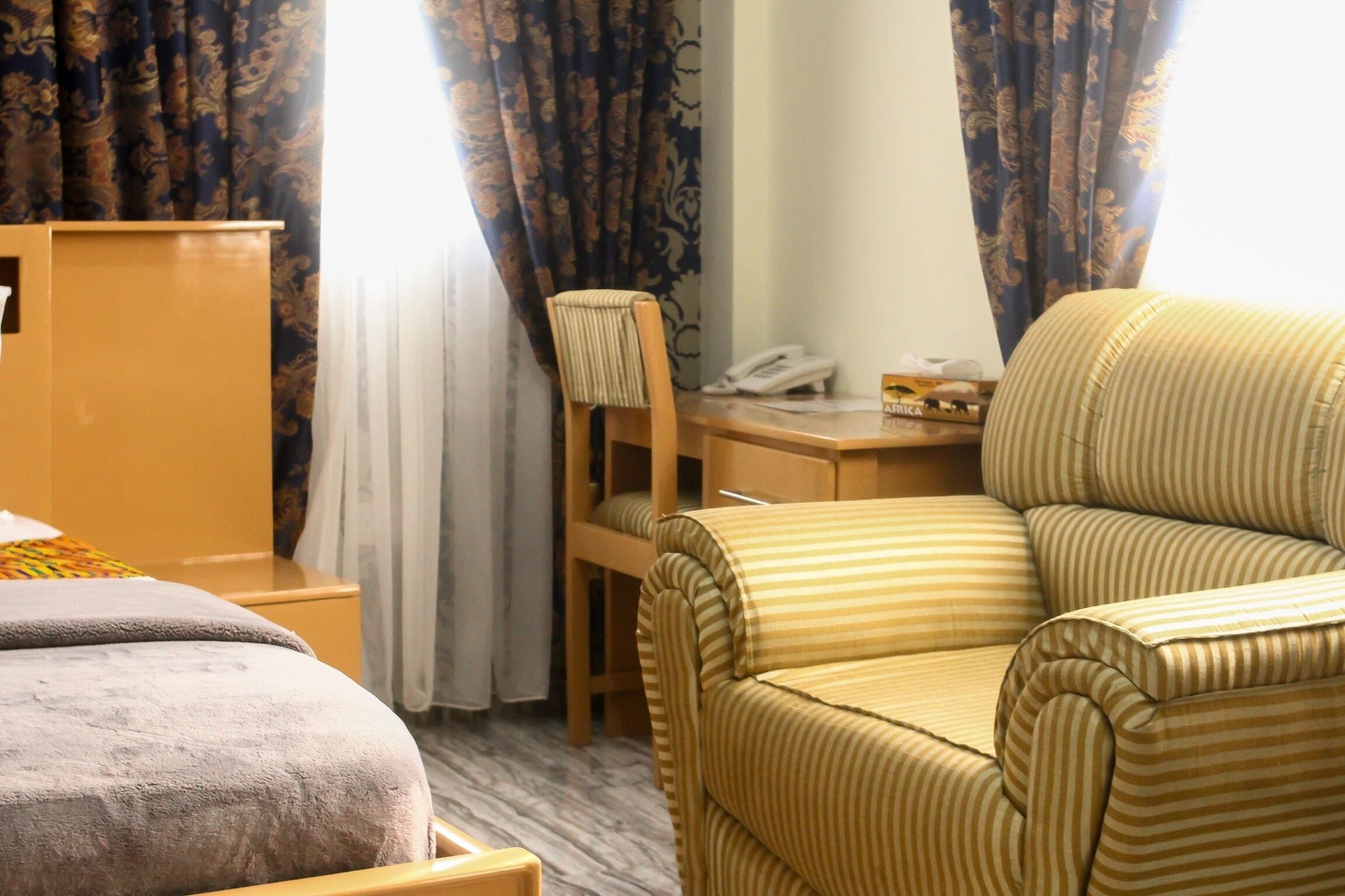 A hotel room with a bed, a striped armchair, a desk with a telephone, and a wardrobe.