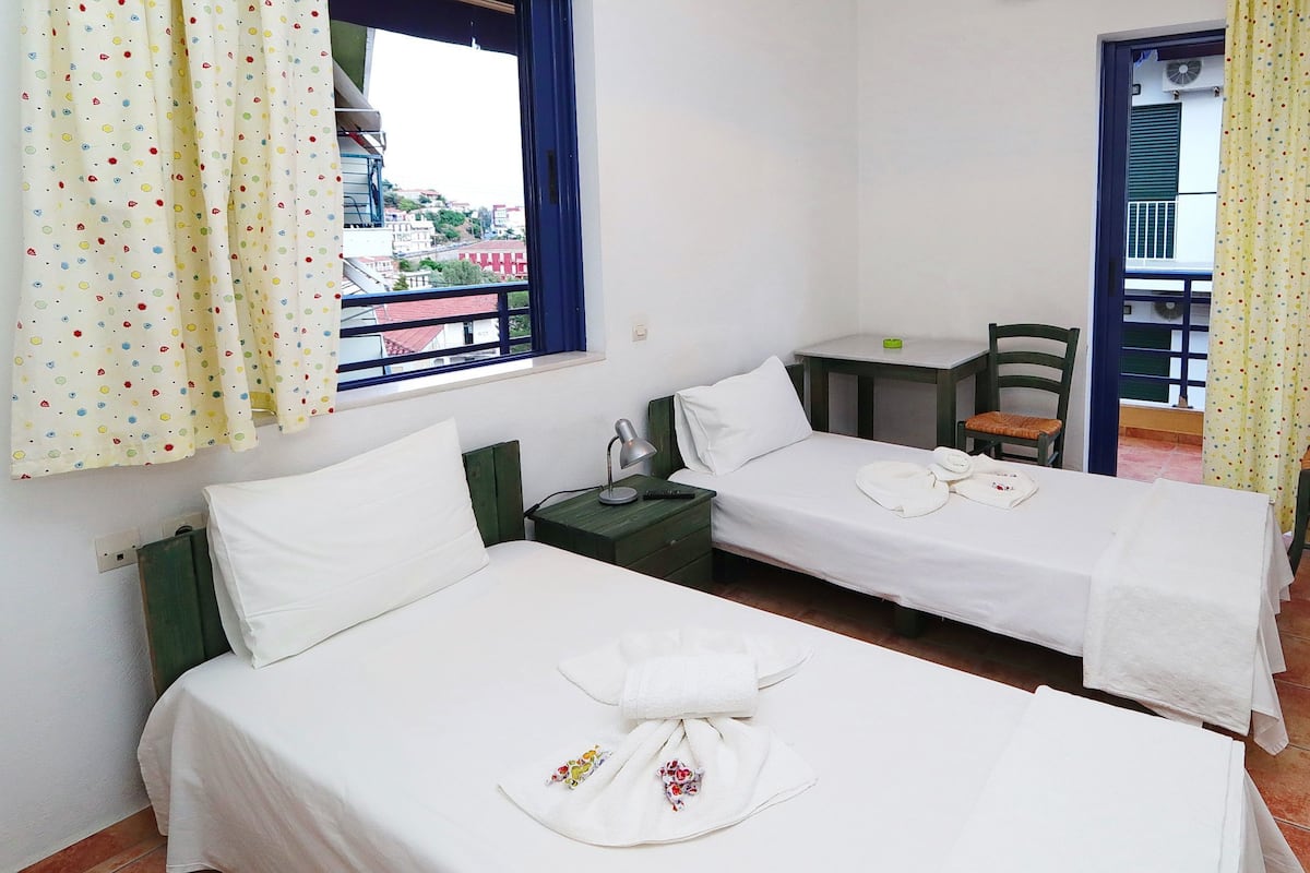 A hotel room with two single beds, a small table, a chair, and a window with curtains.