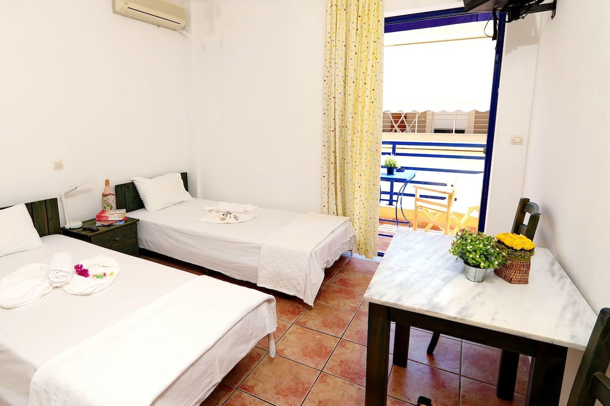 A hotel room with two beds, a small table, a chair, and a balcony with a view.