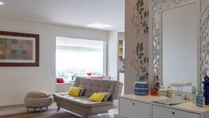 Ein modernes Wohnzimmer mit einer Couch, einem runden Hocker, einer weißen Kommode und einem großen Spiegel, ergänzt durch dekorative Wandbilder.