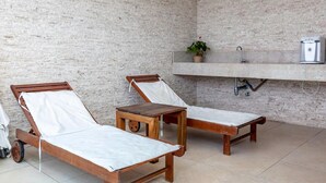 Ein Zimmer mit zwei Holzliegestühlen mit weißen Kissen, einem kleinen Holztisch, einem Blumentopf, einem festinstallierten Spülbecken und einem Wasserspender an der Wand.