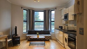 Un espace de vie compact comprenant un canapé, une petite table, un téléviseur, un coin cuisine avec un four à micro-ondes et un lave-linge, ainsi que de grandes fenêtres donnant sur l’extérieur.