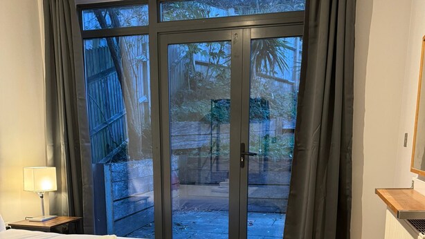 Une chambre à coucher avec un lit, une table de chevet, une lampe, une fenêtre avec des rideaux et une vue sur l’extérieur.