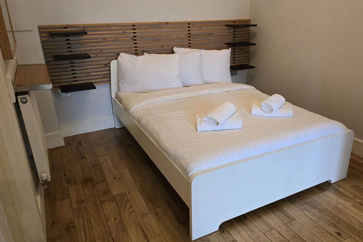 Una cama bien hecha con sábanas blancas y dos toallas enrolladas encima, un cabecero de madera y piso de madera.