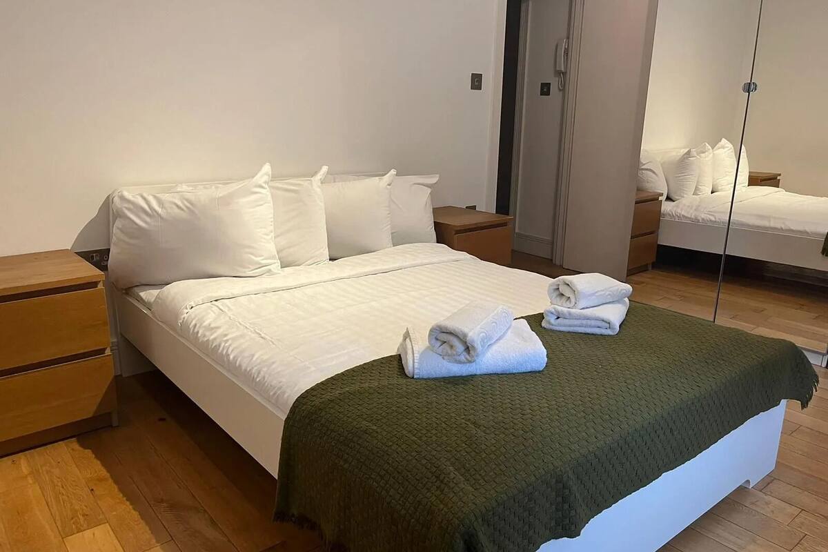 Una cama bien hecha con sábanas blancas y una manta verde, mesitas de noche de madera y un espejo que refleja la habitación.