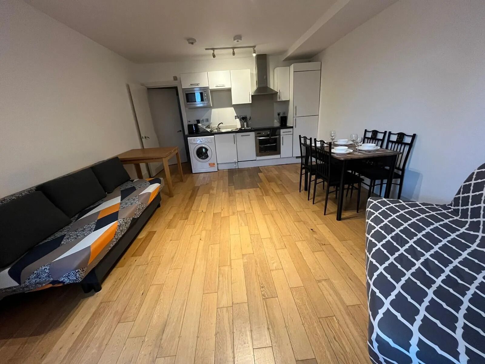 Ett kompakt bostadsutrymme med en soffa, ett matbord med stolar, ett köksområde med vitvaror och en tvättmaskin.