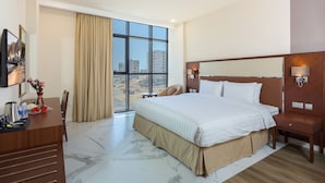 A hotel room with a large bed, a desk with a vase of flowers, a chair, and a large window with a view of buildings. - Muscat Gate Hotel (Muscat)