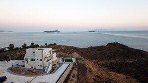 Ein modernes, weißes Gebäude mit Dachterrasse, gelegen an einem felsigen Hang mit Blick auf ein ruhiges Meer und entfernte Inseln.