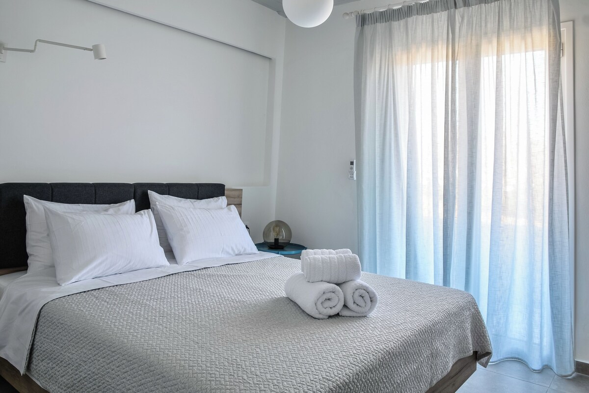 Una cama bien hecha con ropa blanca y almohadas, una mesita de noche con una lámpara y una ventana con cortinas traslúcidas.