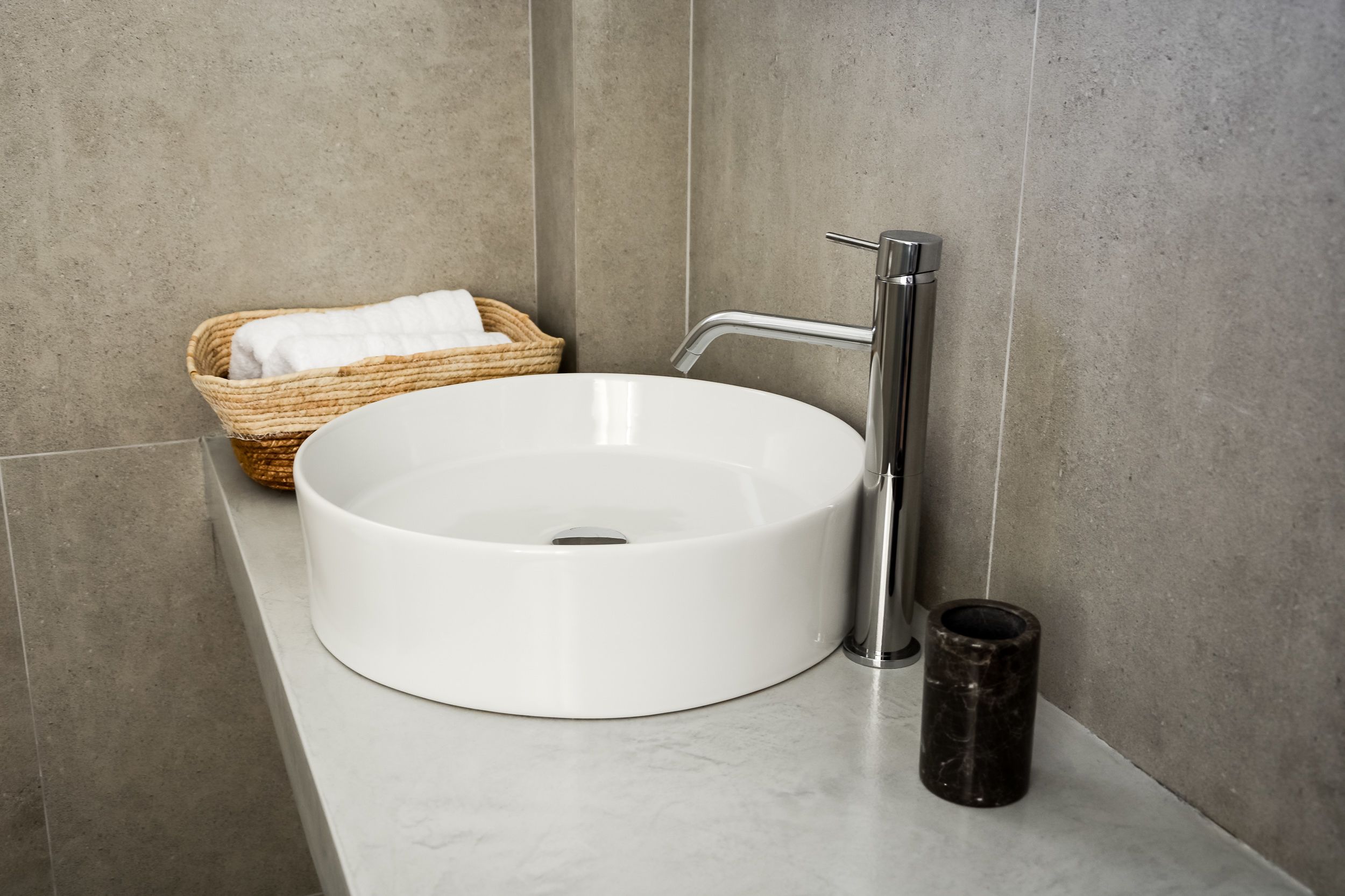 A bathroom with a round white sink, a chrome faucet, a small black vase, and a wicker basket with a white towel.