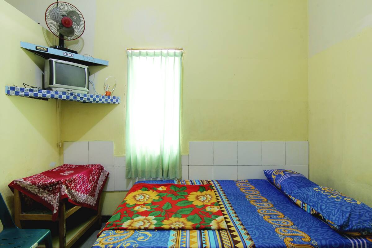 A small, simple room with a bed, a chair, a table with a red tablecloth, a television on a shelf, and a window with curtains.