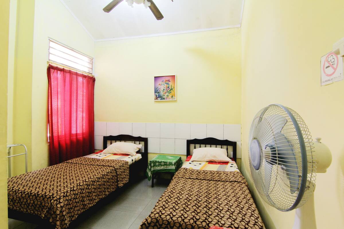 A room with two single beds, a standing fan, a window with red curtains, a small table with a green tablecloth, and a wall-mounted sign.