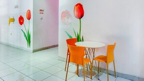 Um corredor com piso de azulejos brancos, uma mesa redonda com cadeiras laranja e uma parede com uma pintura de tulipas vermelhas.
