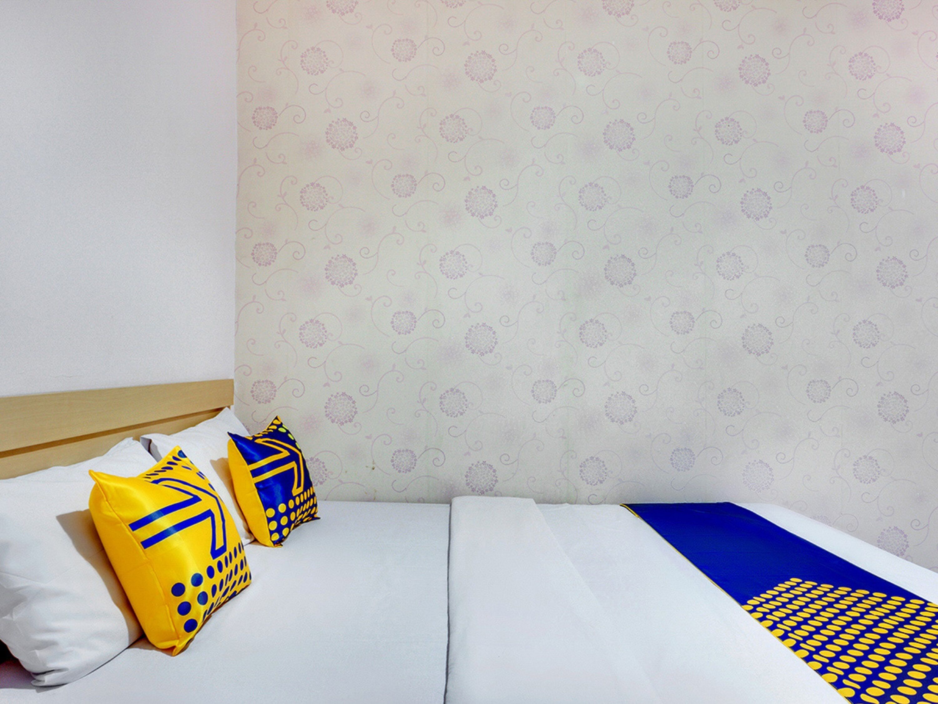 A neatly made bed with white bedding and two pillows, one yellow and one blue, with a patterned wall in the background.