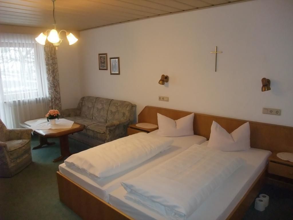 A hotel room with two single beds, a sofa, a small table with a vase, and a cross on the wall.