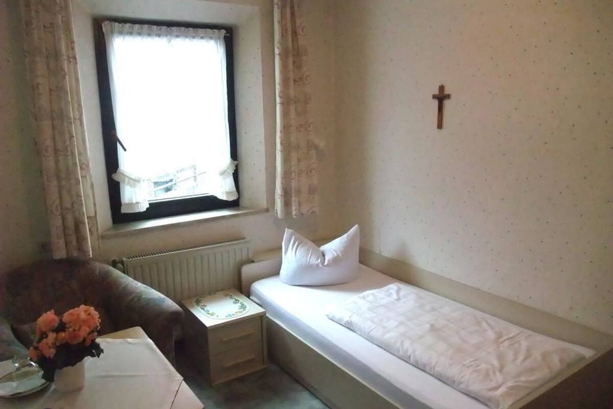 A small, simple room with a single bed, a chair, a small table with a vase of flowers, and a window with curtains.