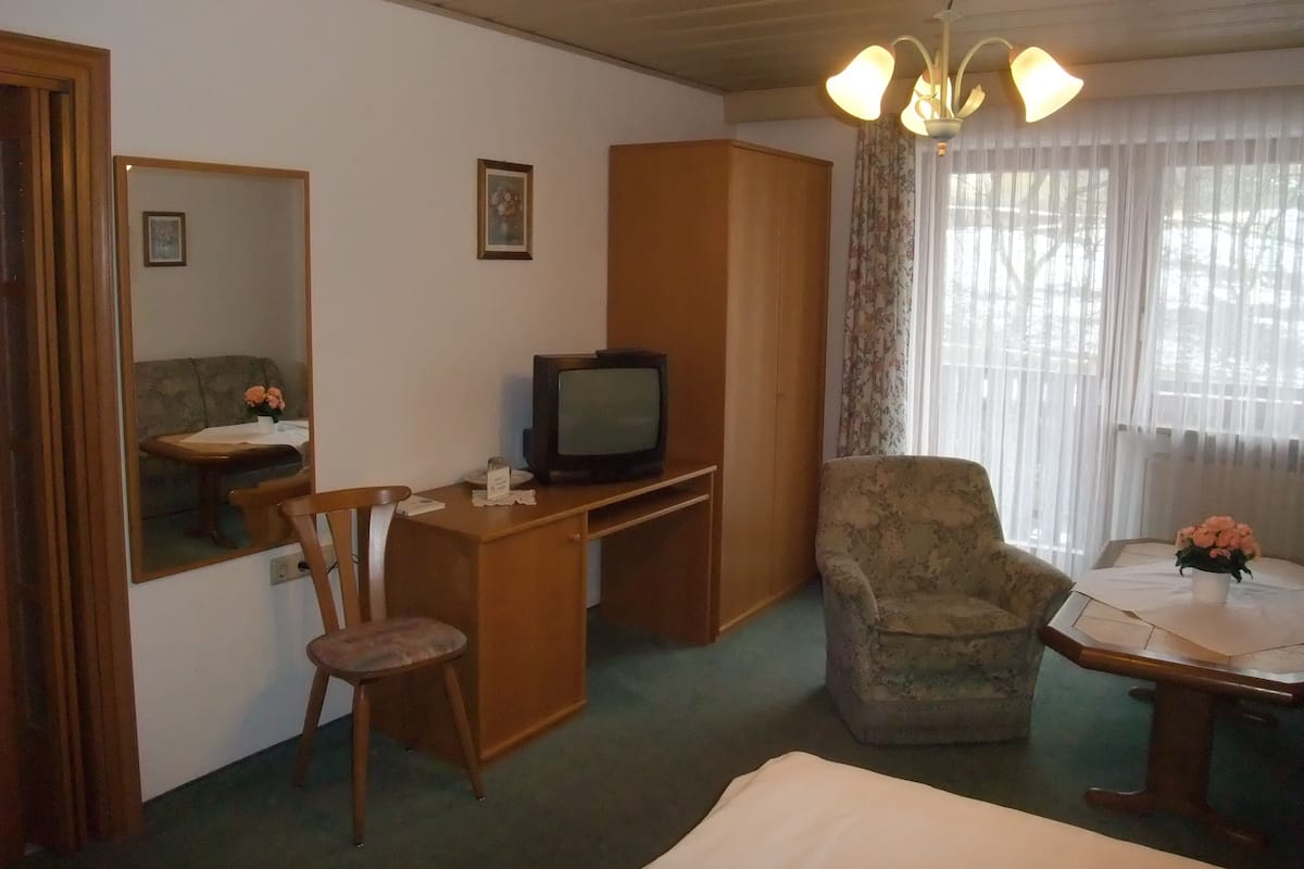A compact hotel room with a wooden desk, a television, a chair, a bed, and a window with curtains.