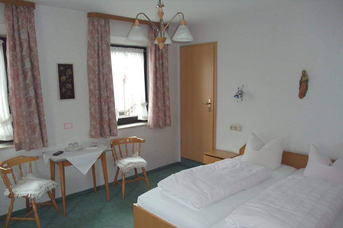 A hotel room with a bed, a desk with a chair, a window with curtains, a door, and a wall-mounted light fixture.
