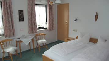 Habitación de hotel con una cama, un escritorio con silla, una ventana con cortinas, una puerta y una lámpara fijada a la pared.