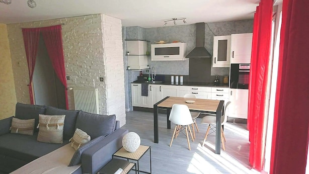 A modern living room with a grey sofa, a small wooden table, and a white lamp. Adjacent to the living room is a kitchen with white cabinets, a microwave, and a red curtain. - Large apartment T2, quiet, near Lyon, balcony parking and transport (Saint-Priest)