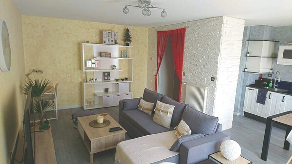 A living room with a sofa, coffee table, and a small side table. There is a bookshelf with various items and a red curtain. A kitchen area is visible in the background.