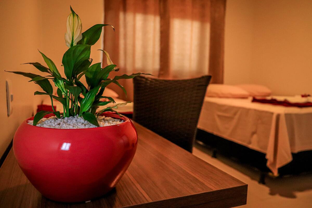 A red pot with green plants on a wooden table in a room with a bed and a chair.