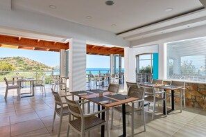 An outdoor dining area with tables and chairs, large windows, and a view of the sea. - Blu Acqua (Agios Nikolaos)