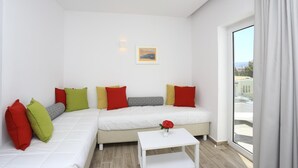 A modern living room with a white sofa, colorful cushions, a small white table with a vase, and a large window offering a city view. - Blu Acqua (Agios Nikolaos)