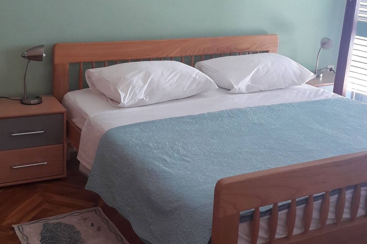 A bedroom with a wooden bed, white pillows, a blue blanket, a nightstand with a lamp, and a wooden floor.