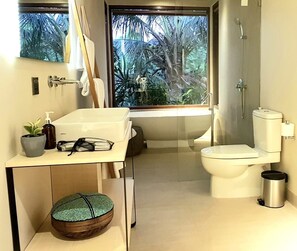 A modern bathroom with a toilet, bidet, and shower. There is a window with a view of palm trees. A small table with a plant and a bottle of soap is visible. - Lin Ubud (Ubud)