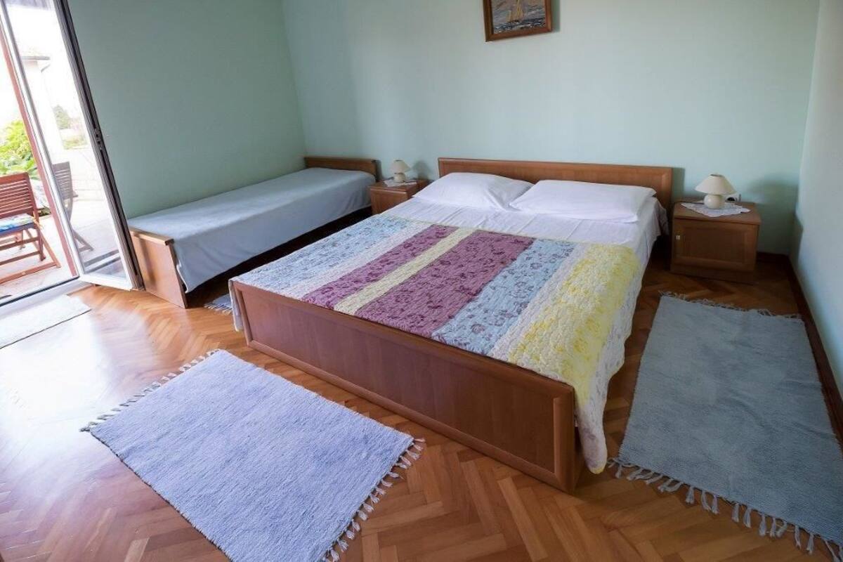 A bedroom with a wooden bed, a smaller bed, two bedside tables with lamps, and a view of the outdoors through a glass door.