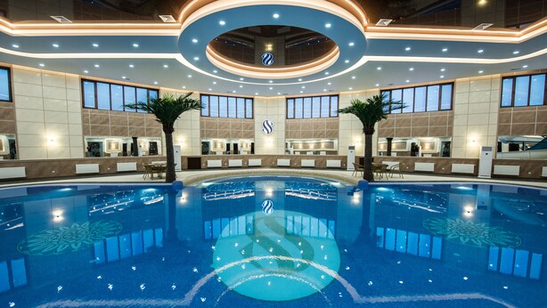 A large indoor swimming pool with a central island, surrounded by reception desks and palm trees, and a modern ceiling design. - Simma Hotel spa and waterpark (Tashkent)