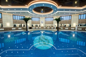 Une grande piscine intérieure dotée d’un îlot central, entourée de comptoirs d’accueil et de palmiers, et agrémentée d’un design de plafond moderne.