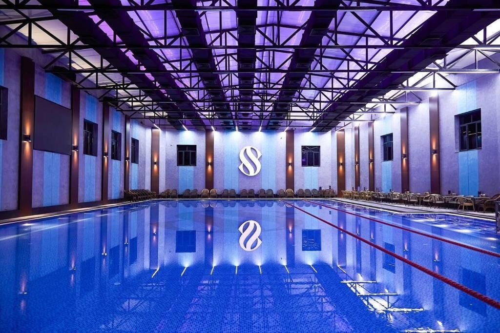 A large indoor swimming pool with a modern design, featuring a geometric ceiling structure and a series of windows along the walls.
