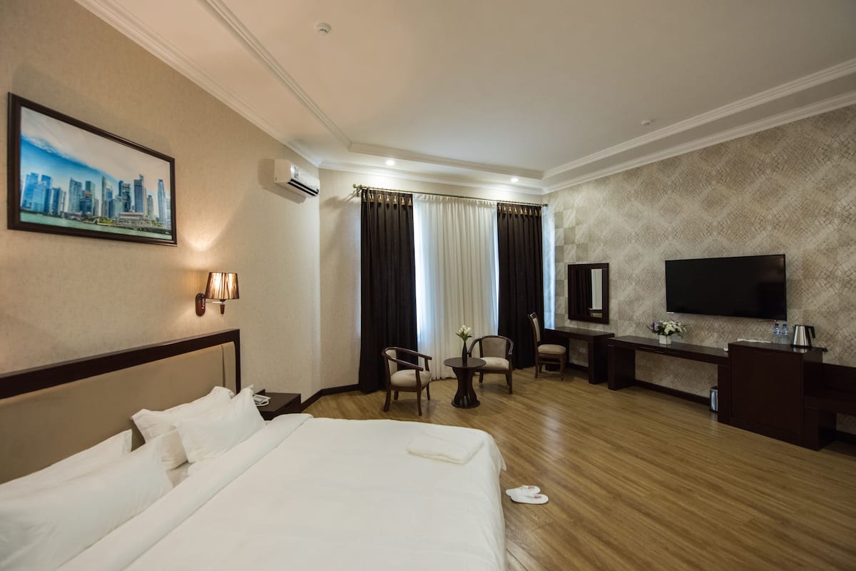 Quarto de hotel com uma cama grande, uma escrivaninha com televisão de tela plana, um espaço de estar com cadeiras e uma pintura da paisagem urbana na parede.