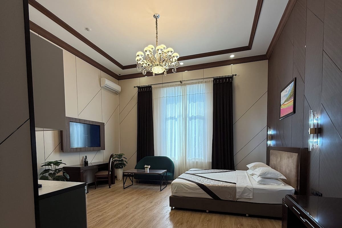 Quarto de hotel moderno com piso de madeira, uma cama, uma escrivaninha com cadeira, televisão e lustre.