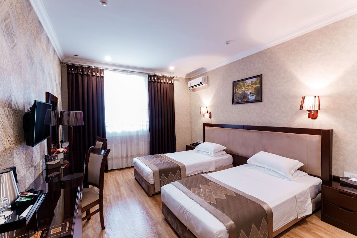 Quarto de hotel com duas camas, uma escrivaninha com cadeira, televisão e uma janela com cortinas.