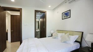 A bedroom with a large bed, a bedside table with a lamp, a wall-mounted picture, and a door leading to another room. - Goa Junction Vagator - Apparthotel (Vagator)