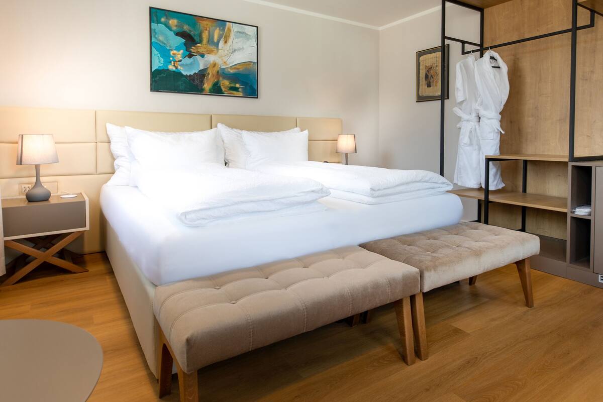 A hotel room with a bed, bedside tables, a bench, a robe rack, and a painting on the wall.