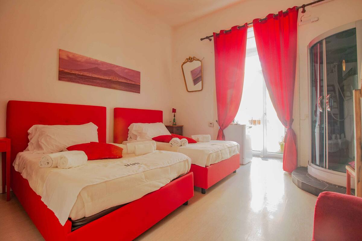 A hotel room with two beds, red curtains, a large window, and a painting on the wall.