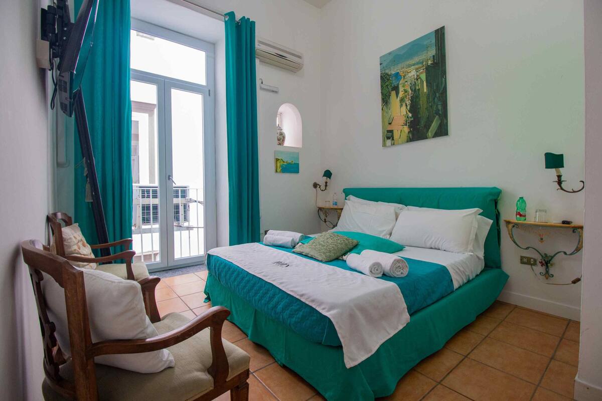 A bedroom with a bed, a chair, a window with curtains, a painting, and a wall-mounted shelf.