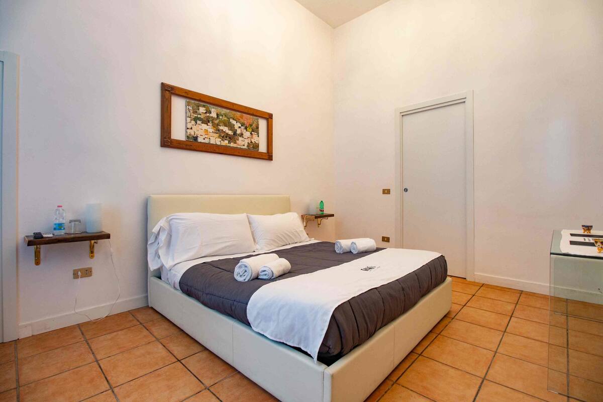 A neatly made bed with a white and brown bedspread, a small bedside table with a lamp, and a framed picture on the wall.