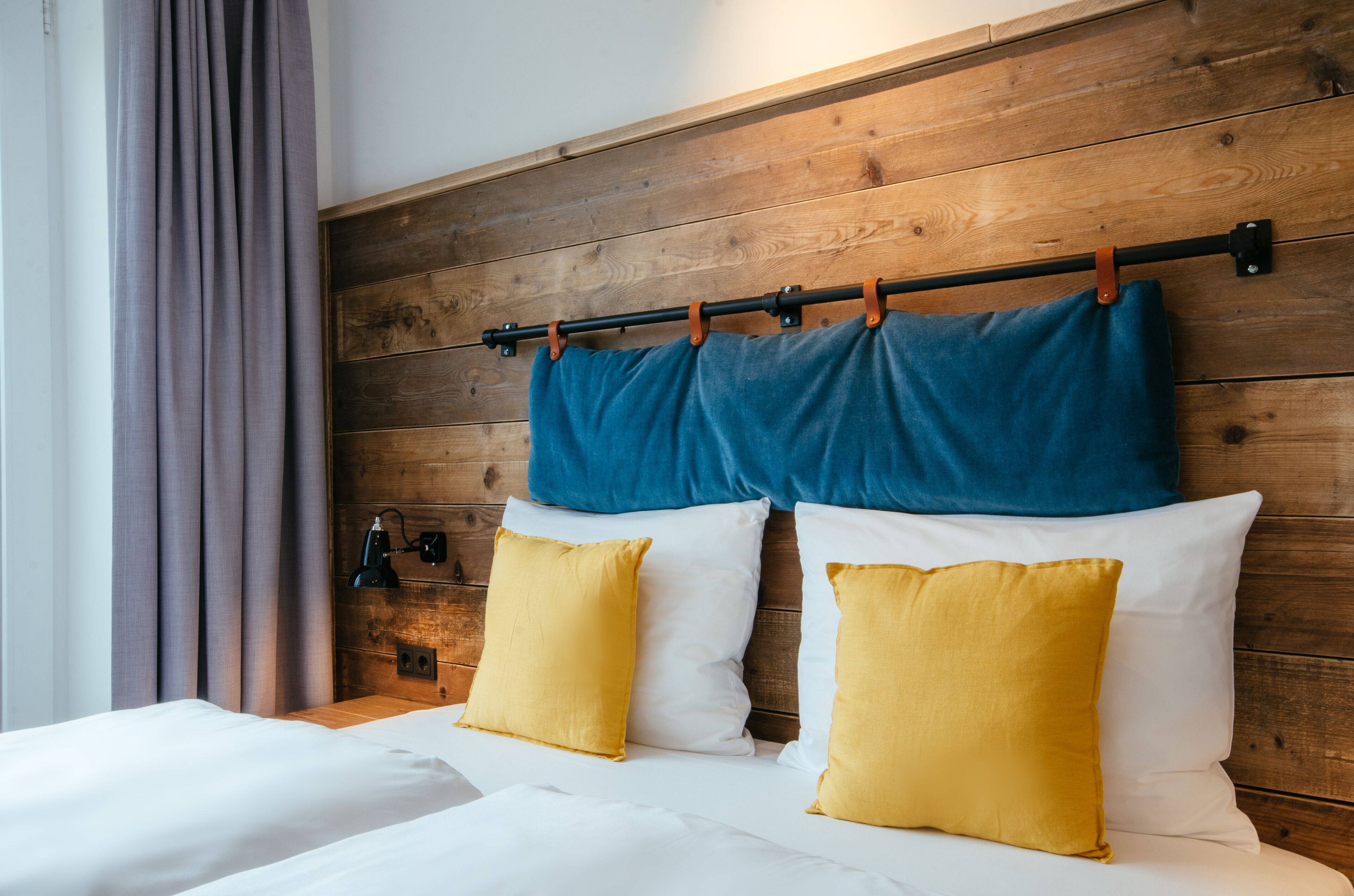 A hotel room with a wooden headboard, two beds with white and yellow bedding, and a window with curtains.