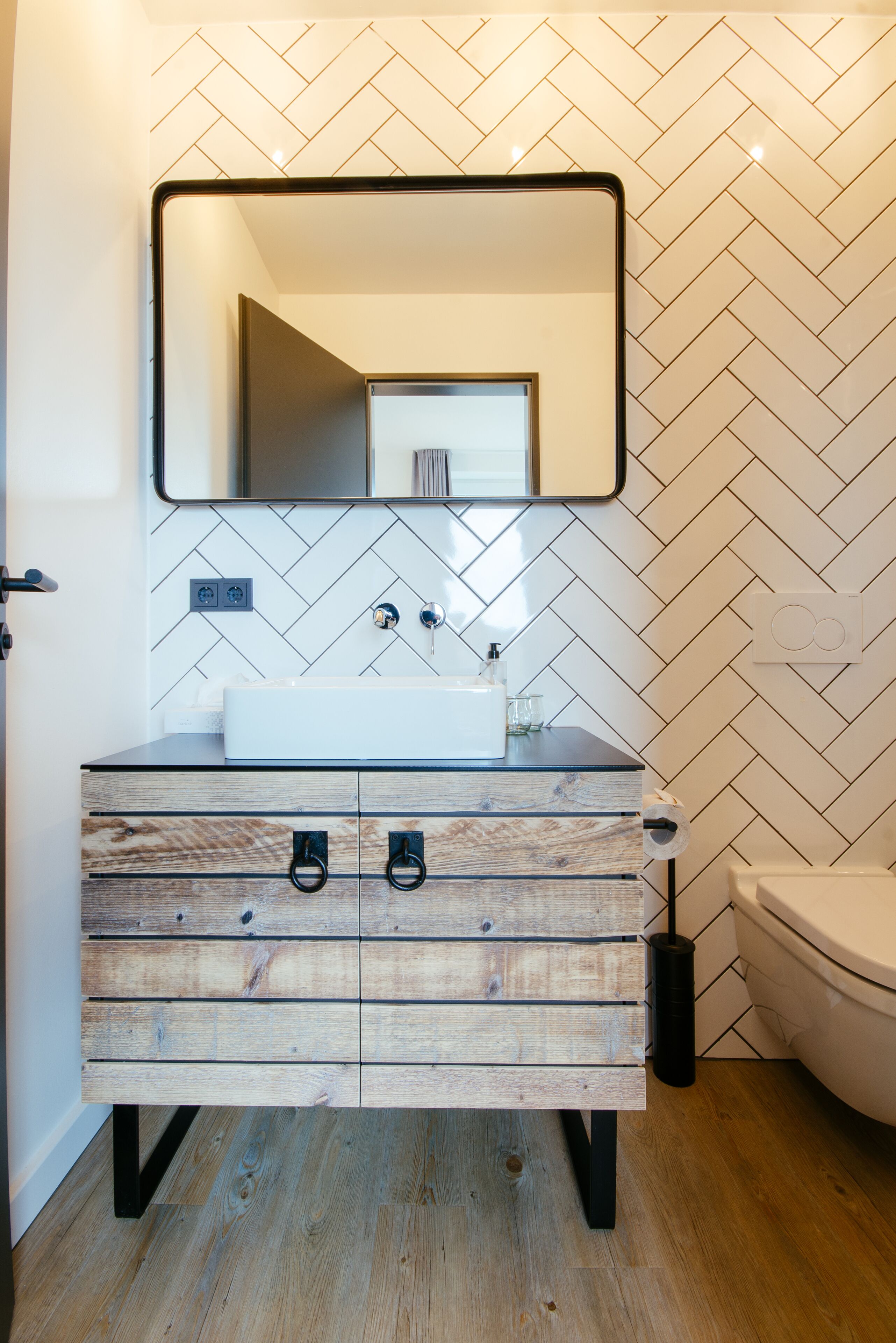 A bathroom with a white herringbone-patterned wall, a wooden vanity with drawers, a mirror, and a toilet.