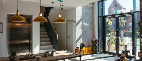 A modern interior with a wooden reception desk, a staircase, and large windows offering a view of the outside.