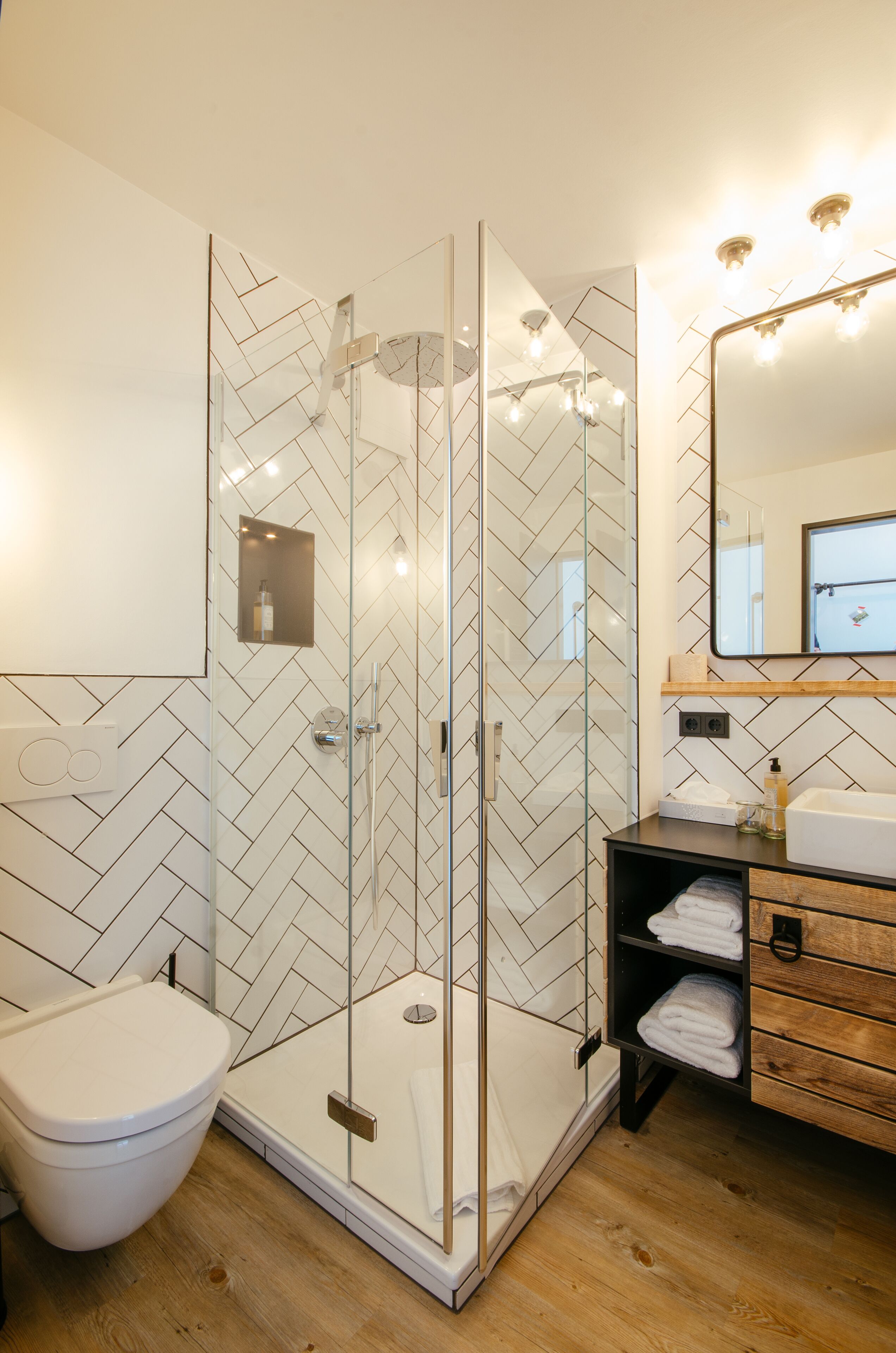 A modern bathroom with a glass-enclosed shower, a white toilet, and a wooden vanity with a mirror and shelves.