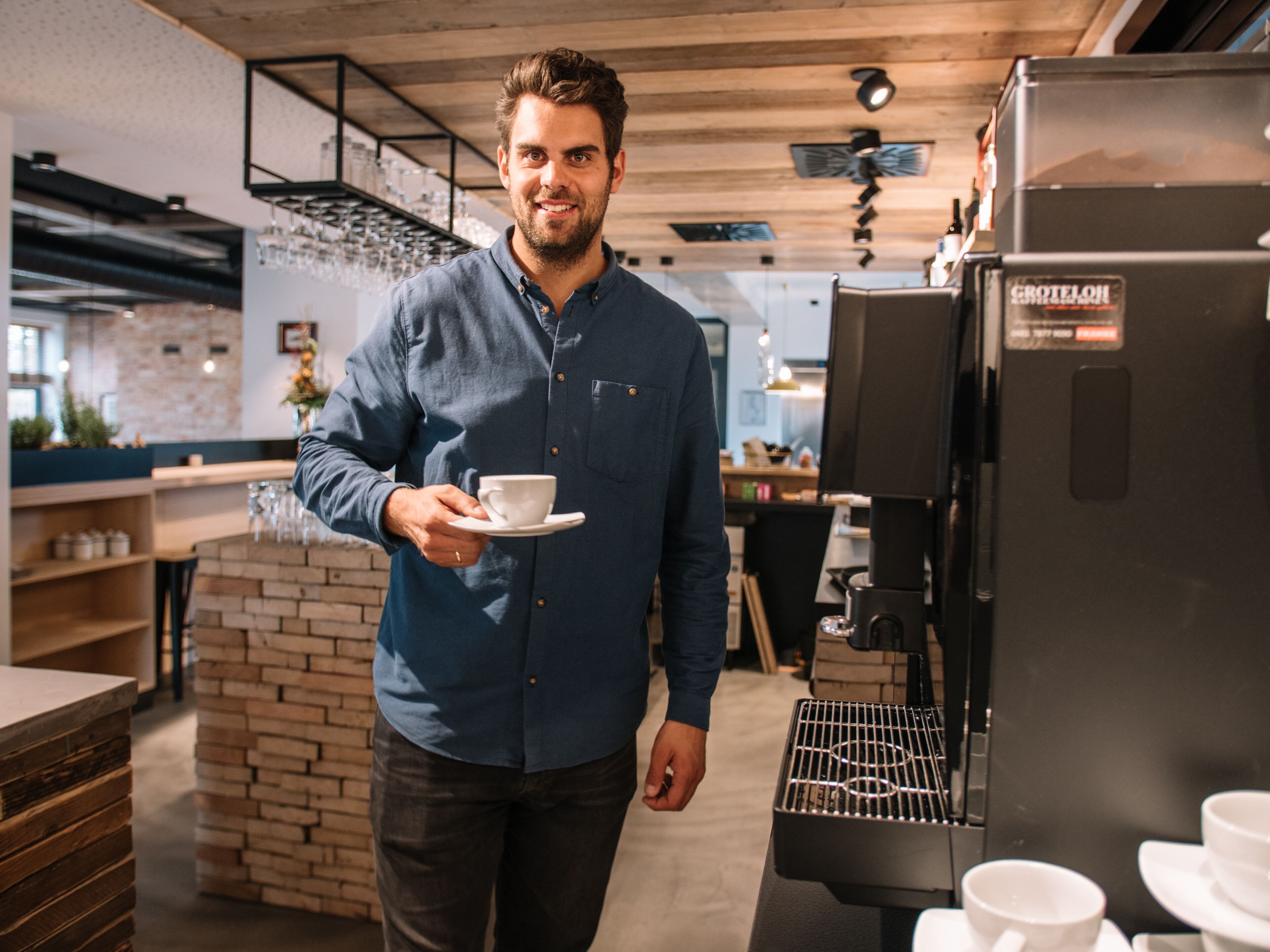 A person in a blue shirt holding a cup, standing in a modern kitchen with a coffee machine and brick wall.