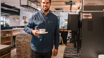 A person in a blue shirt holding a cup, standing in a modern kitchen with a coffee machine and brick wall.